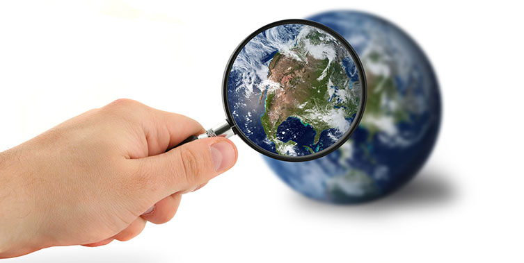 A person's hand holds a magnifying glass in front of a globe, making one of the continents appear larger.